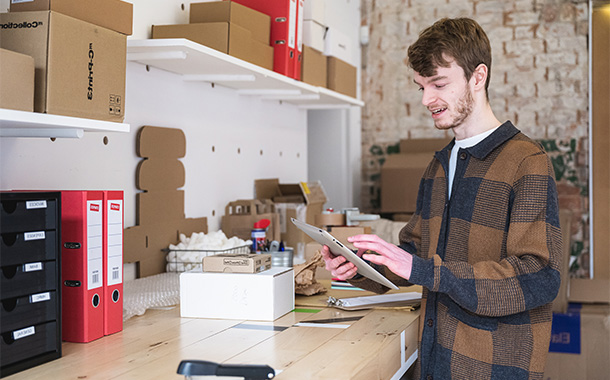 Person using a tablet to manage business operations in a small warehouse with shelves, boxes, and packing materials