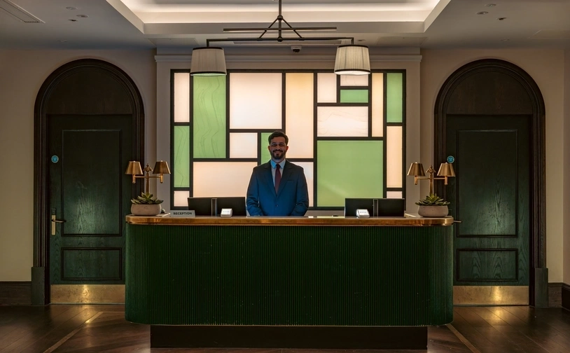 A hotel reception desk with a suited staff member standing behind a dark green counter, set in an elegant lobby with geometric glass panels, brass lamps, and arched doorways.