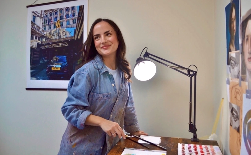 Artist Elysia Gilman works at a table in her studio in North Wales, surrounded by sketches, reference images and painting tools, with a desk lamp illuminating her workspace