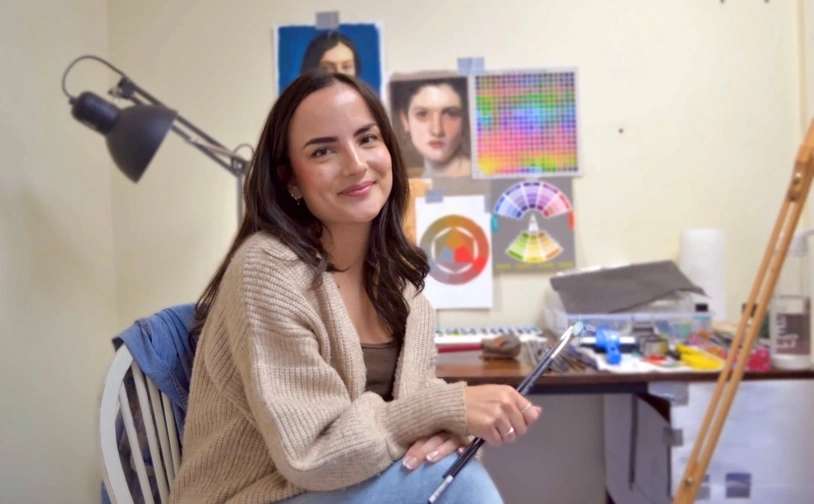 Artist Elysia Gilman sits in her studio in North Wales, holding a paintbrush, with sketches, colour charts and art materials arranged on the wall and worktable behind her