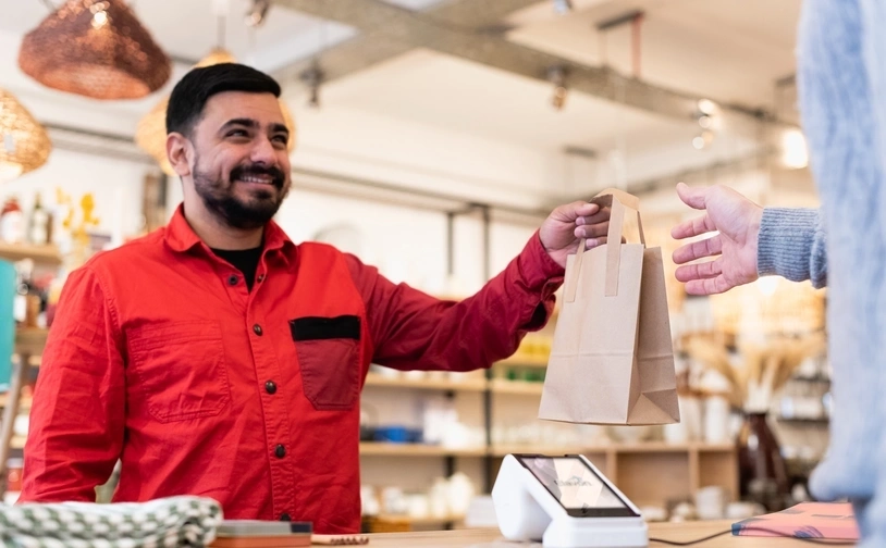 A retail employee hands a paper shopping bag to a customer across the counter, with a payment terminal visible in the foreground