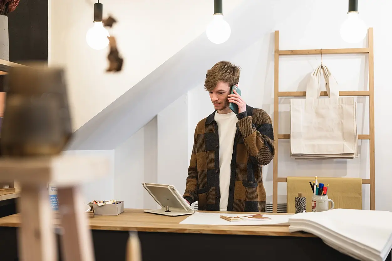 Person standing behind a wooden counter in a modern shop, speaking on a phone and using a tablet, with shelves and hanging tote bags in the background.