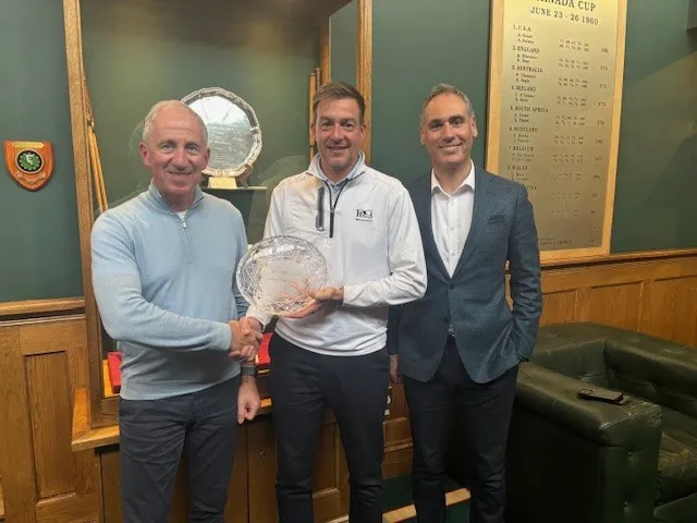 Three people indoors presenting a crystal trophy at a golf awards ceremony, with a wooden honours board and shield in the background.